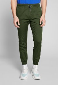 Pantalon de jogging vert avec une taille élastique et un cordon de serrage ajustable ; coupe effilée, texture lisse et poignets élastiques. Associé à des baskets.