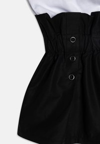 Černá sukně z látky se shromážděným pasem. Obsahuje stříbrné očky a hladkou texturu. Součástí je bílá horní část s krátkými rukávy.