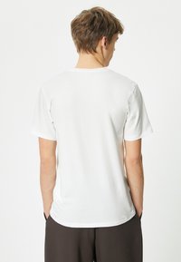 Witte t-shirt met korte mouwen, gemaakt van zachte stof. Klassieke ronde hals, eenvoudig ontwerp en relaxte pasvorm, gedragen met de handen in de zakken van donkere broek.