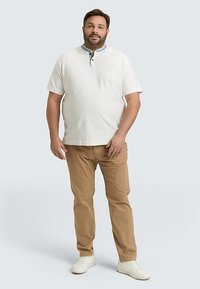 Wit poloshirt met korte mouwen, een kraag en knoopdetails, gecombineerd met lichtbruine broek en witte sneakers. Effen achtergrond.
