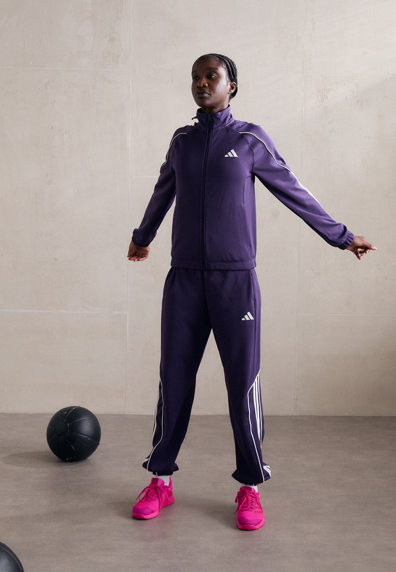 Violetinės sportinės treniruočių kelnės su baltais dryžiais, su užtrauktuku užsegama striuke ir elastingomis kelnėmis. Ryškiai rožiniai sportiniai bateliai užbaigia aprangą.