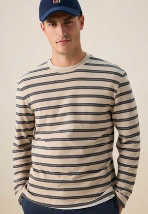 Jonge man in een beige en marineblauw gestreept shirt met lange mouwen en een marineblauw pet met een kleurrijk logo, handen in de zakken, neutrale achtergrond.