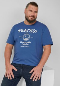 Niebieska bawełniana koszulka z krótkim rękawem i okrągłym dekoltem z białym nadrukiem "Field Trip" oraz grafiką. Połączona z ciemnymi dżinsami.