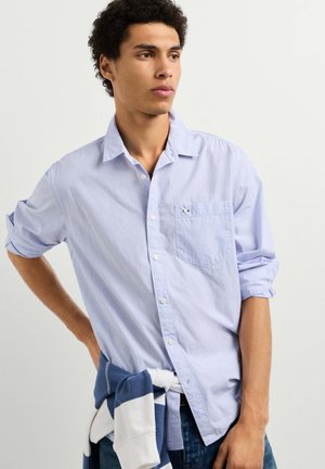 Jonge man die een lichtblauw hemd met knopen en opgerolde mouwen draagt, en een blauw-witte trui om zijn middel geknoopt heeft.
