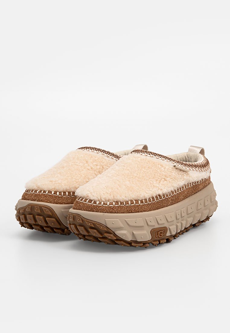 Zapatos beiges tipo slip-on con parte superior suave y peluda, entresuela marrón texturada y suela robusta con un patrón de panal y detalle de logo.