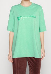 Osoba oblečená v volném mátově zeleném tričku s logem "KARL LAGERFELD JEANS" v zelené a bílé na přední straně, doplněném o tmavě červené sametové kalhoty.