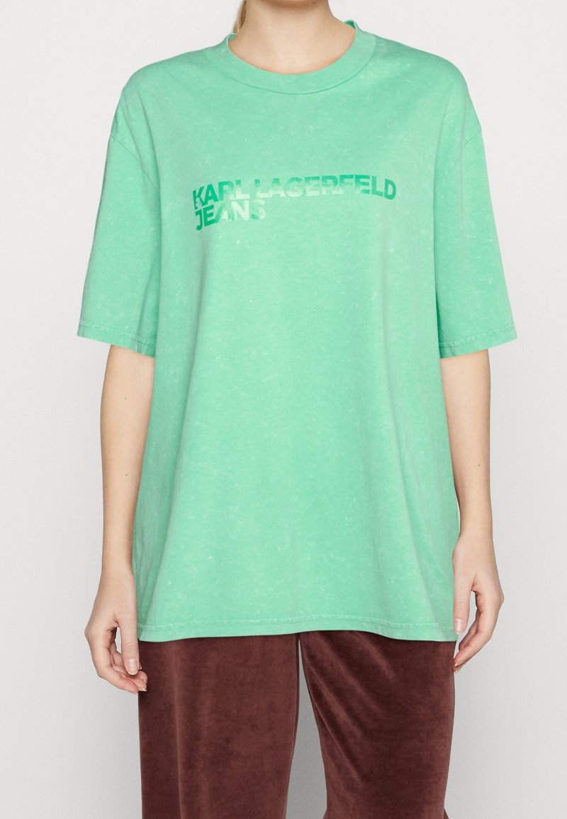 Osoba oblečená v volném mátově zeleném tričku s logem "KARL LAGERFELD JEANS" v zelené a bílé na přední straně, doplněném o tmavě červené sametové kalhoty.