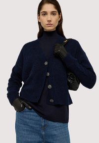 Námořnický modrý pletený kardigan s texturovaným vzorem, pět knoflíků vpředu, spárovaný s černou kabelkou a koženými rukavicemi.