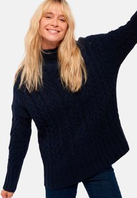 Pull en maille côtelée bleu marine avec un col haut, des épaules tombantes et une coupe ample. L'ourlet côtelé et les larges poignets ajoutent une touche de texture.