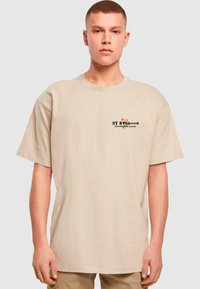 Beige oversized t-skjorte i bomull med korte ermer, med et svart og flerfarget grafisk logo på brystet. Enkel, avslappet design.