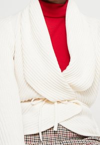 Cárdigan de punto acanalado en color crema con amplias solapas, cintura ceñida y tejido texturizado. Lleva debajo un cuello alto rojo. Un tejido a cuadros asoma por debajo.