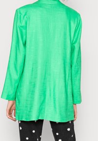 Blazer verde con un brillo texturizado, con mangas largas y cuello de solapa, combinado con pantalones negros decorados con patrones florales blancos.