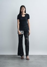 Nő fekete, rövid ujjú, kulcslyuk kivágásos felsőben, fekete, csipkés, bő szárú nadrágban, fekete magassarkúban, szürke borítéktáskával a kezében, egy egyszerű fal előtt áll.