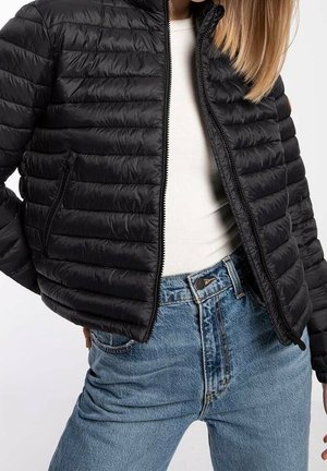 Personne portant une veste matelassée noire sur un haut blanc et un jean bleu taille haute sur un fond uni.