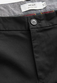 Schwarze, enganliegende Hose mit glatter Textur, ausgestattet mit einem Knopfverschluss und einem grauen gemusterten Futter. Markenlabel sichtbar im Taillenbund.