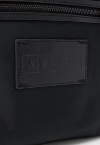 Black nylon bag featuring a textured leather patch labeled "AX / ARMANI EXCHANGE". Zipper closure along the top, minimalist design, no patterns.