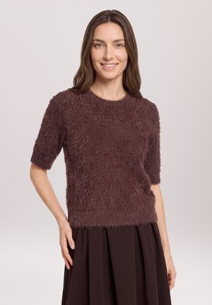 Femme souriante aux longs cheveux bruns porte un pull marron duveteux à manches courtes et une jupe plissée marron, debout devant un fond clair uni.