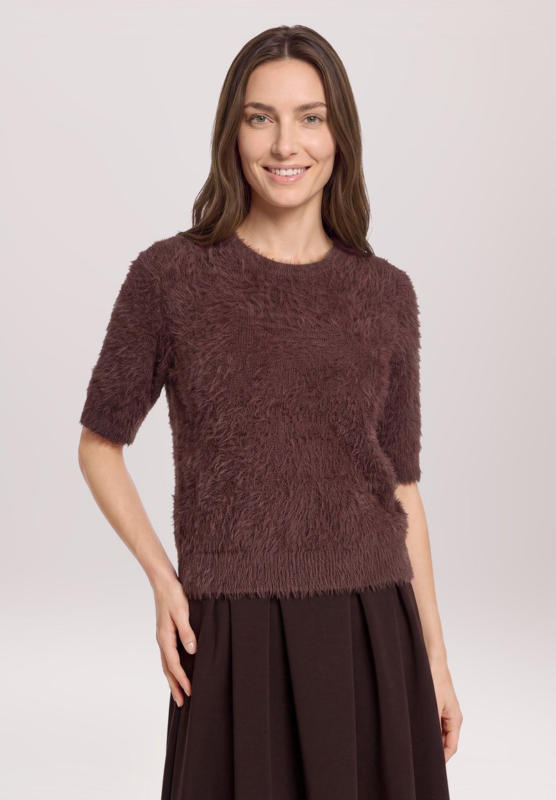 Femme souriante aux longs cheveux bruns porte un pull marron duveteux à manches courtes et une jupe plissée marron, debout devant un fond clair uni.
