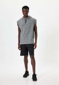 Sudadera sin mangas gris con un bolsillo y cordón, combinada con pantalones cortos negros. El modelo lleva zapatillas deportivas negras. Texturas suaves en todo.