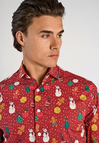 Rode overhemd met knopen, versierd met witte stippen en een feestelijk patroon van sneeuwpoppen, ornamenten en peperkoeken figuren in verschillende kleuren.