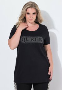 Mujer vestida con una camiseta negra con la palabra "QUEEN" en pedrería, posando con una mano cerca de su cuello y leggins con franjas laterales de estampado de leopardo.