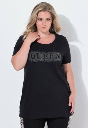 Žena v černém tričku s nápisem "QUEEN" vyloženým kamínky, pózuje s jednou rukou u krku a legínami se zvířecím potiskem po stranách.