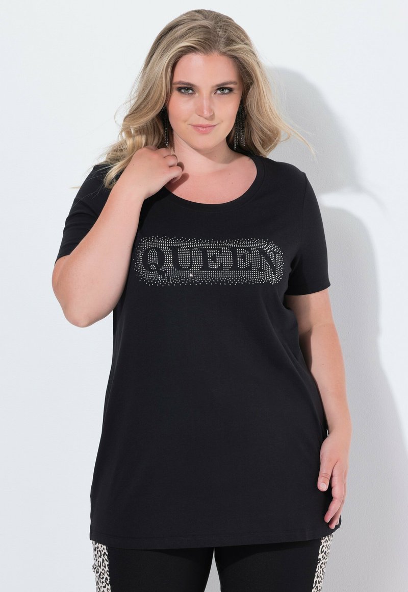 Mujer vestida con una camiseta negra con la palabra "QUEEN" en pedrería, posando con una mano cerca de su cuello y leggins con franjas laterales de estampado de leopardo.