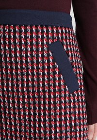 Kudottu ruudullinen hame, jossa on punaisen, valkoisen ja laivastonsinisen sävyjä. Sisältää laivastonsinisen vyötärönauhan ja sivutaskun, jossa on laivastonsininen kangasdetaili.