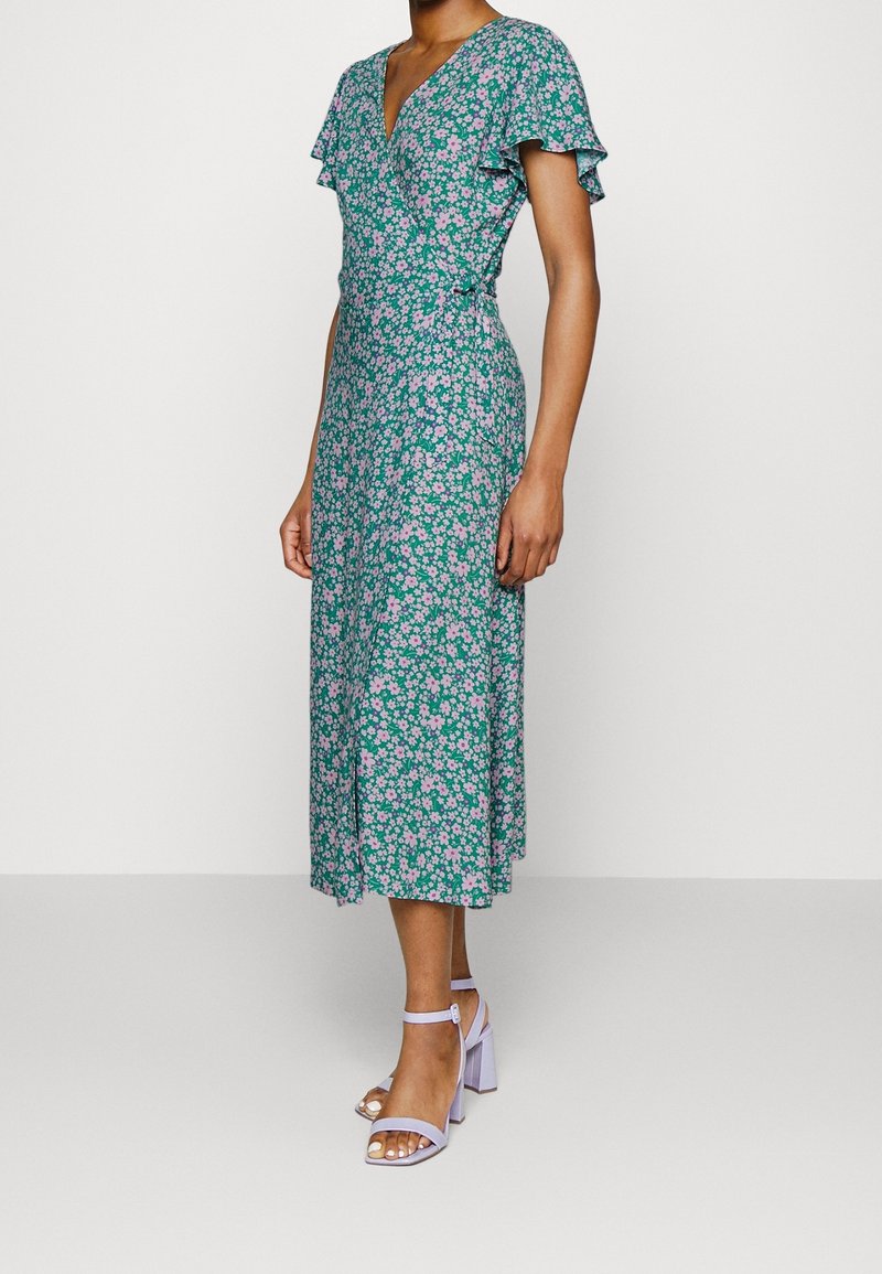 Robe florale verte avec des accents roses, manches courtes et flottantes, design enveloppant et longueur midi. Associée à des sandales à talons carrés lavande.