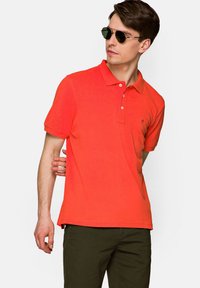 Czerwony polo z klasycznym kołnierzykiem, trzema guzikami, krótkimi rękawami i haftowanym logo po lewej stronie klatki piersiowej. Noszone z ciemnozielonymi spodniami.