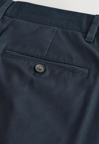 Dunkel marineblaue Hose mit einer hinteren Tasche, die mit einem einzelnen Knopf verschlossen wird, und einer Gürtelschlaufe darüber, die Stoffstruktur und Nahtdetails zeigt.