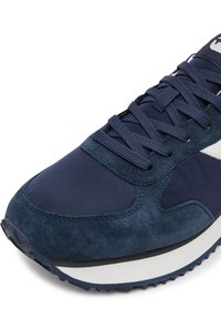 Naktaudu zilas sporta kurpes ar zamšāda un neilona materiālu kombināciju, teksturētām sānu daļām, plakaniem aukliņām un baltu gumijas zolīti.