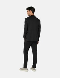 Homme debout, de dos, portant un costume gris foncé avec un pantalon assorti et des baskets gris clair sur un fond blanc uni.