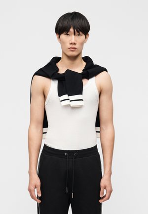 Young man with short black hair wearing a white sleeveless top, black sweatpants, and a black sweater draped over his shoulders.