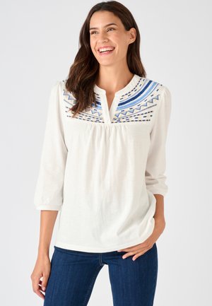 Witte blouse met ingewikkeld blauw borduurwerk rond de halslijn en driekwart mouwen, gecombineerd met donkerblauwe spijkerbroek. Zachte stoftextuur.