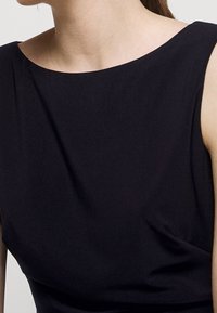 Žena v černých šatech bez rukávů se lodičkovým výstřihem, který odhaluje její krk, ramena a horní část hrudníku.