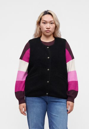 Jeune personne aux longs cheveux blonds portant un gilet noir boutonné sur un pull à rayures rose, blanc et violet à manches longues et un jean bleu.