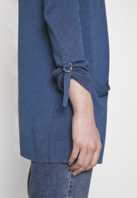 Modrý pletený svetr s volným střihem, s žebrovanými manžetami a dekorativním kroužkem na rukávu. Materiál vypadá měkce a strukturovaně.