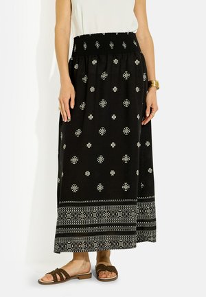 Jupe maxi noire à motifs géométriques blancs, taille élastique, portée avec un top blanc, des sandales slides marron, et des bracelets et bagues en or.