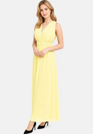 Donna in piedi che indossa un vestito lungo senza maniche giallo pallido con vita stretta e scarpe nere con tacchi alti su sfondo bianco.