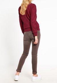 Žena v burgundském svetr, hnědých slim-fit kalhotách s ohrnutými kotníky a bílých sklápcích, stojící na bílem pozadí.