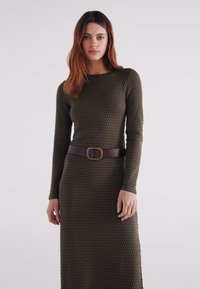 Olijfgroene jurk met lange mouwen, gestructureerde stof met een luchtbelpatroon, strakker bij de taille met een bruine riem en een decoratieve gesp.