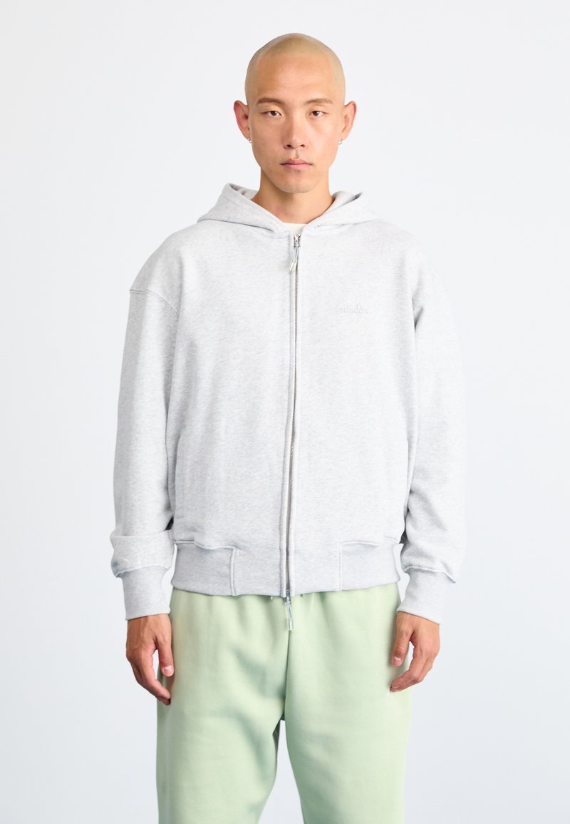 Siv žametni hoodie na zadrgo z kapuco, elastičnimi manžetami in robom. Nosen preko svetlo zelenih trenirk, ki imajo sprednje žepe in sproščen kroj.