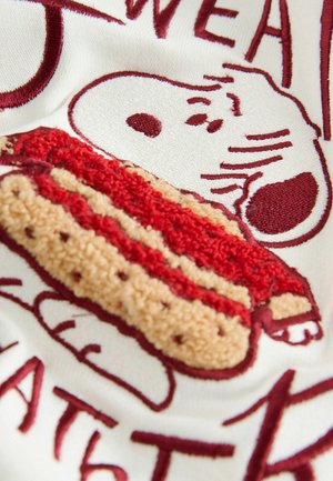 Vyšívaná bílá tkanina obsahuje stylizovaného Snoopyho a párek v rohlíku, s červenými a béžovými akcenty, vytvářejícími texturovaný a hravý design.