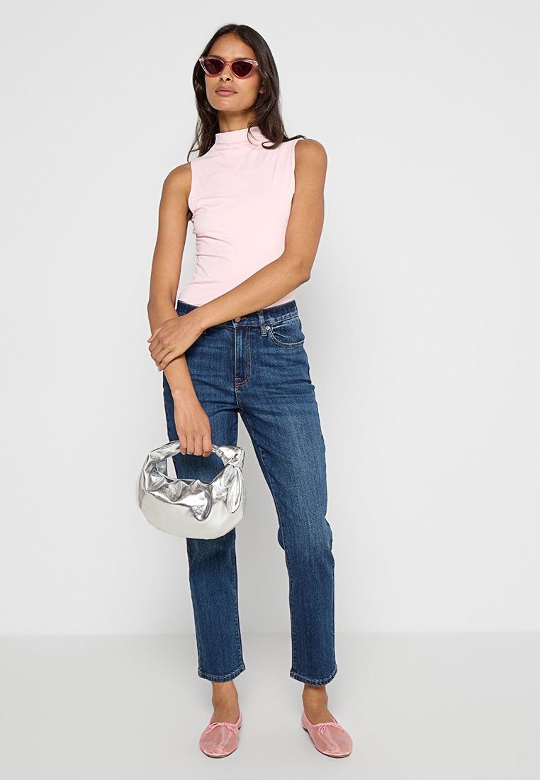 Rosa ärmelloses Oberteil mit Stehkragen, blaue gerade geschnittene Jeans, silberne strukturierte Handtasche und rosa Slipper. Eine Sonnenbrille vervollständigt das Outfit.
