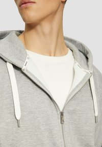Grijze zip-up hoodie met witte trekkoorden, met een zachte textuur en een metalen rits. Draagt over een witte t-shirt met een ronde hals.