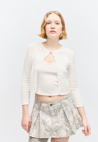 Jeune femme aux cheveux blonds courts portant un cardigan blanc à motifs, un débardeur crème et une jupe plissée gris clair sur un fond uni.