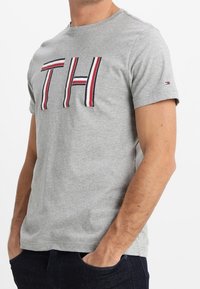 T-shirt en coton gris avec un col rond, des manches courtes et un logo "TH" bien visible, agrémenté de détails à rayures rouges et bleues.