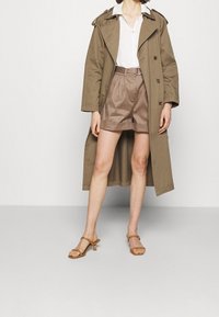 Vrouw in een beige trenchcoat over een witte blouse, met hoog-wijde bruine shorts en tan gekleurde haksandalen, staande op een effen lichte achtergrond.