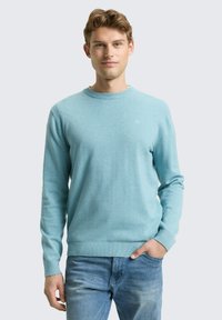 Maglione azzurro chiaro in morbido tessuto, scollo rotondo, maniche lunghe, polsini e orlo a coste, abbinato a jeans blu. Logo discreto sul petto.